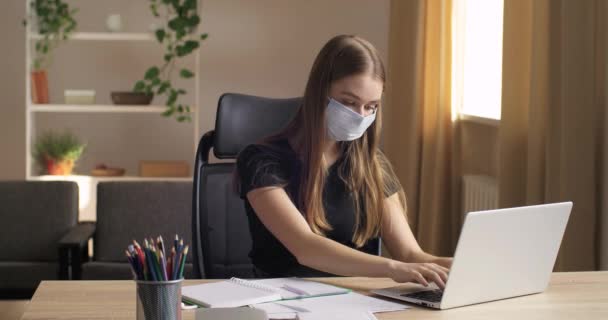Porträt von jungen Teenager-Mädchen Geschäftsfrau trägt medizinische Schutzmaske auf dem Gesicht sitzt im Büro oder zu Hause hinter Laptop arbeitet Online-Drucke auf dem Computer macht Notizen, Fernlernkonzept — Stockvideo