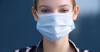 Koruyucu tıbbi bir maske takmış bir kadının yakın plan portresi. Sokakta tek başına duran genç bir kız mavi gözlerle kameraya bakıyor. Üzücü bir bakış, koronavirüs salgını tehlikesi.