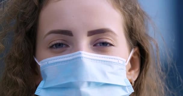 Detailní portrét roztomilé kudrnaté ženy v ochranné masce při pohledu do kamery s modrýma očima, ženský obličej se zavřeným nosem a ústy, prevence pandemické epidemie koronavirového onemocnění — Stock video