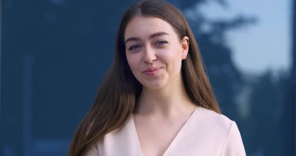 Portrait de jeune étudiante attrayante femme d'affaires aux cheveux longs et aux yeux bleus pose devant la caméra à l'extérieur sur fond de bâtiment de la ville souriant mignon et sympathique embarrassé — Video