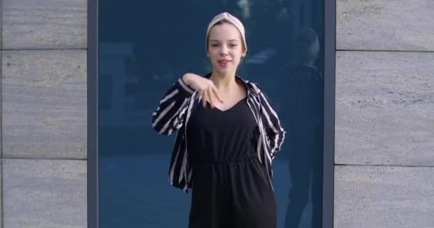 Jeune étudiante active joyeuse porte bandeau et veste rayée bouge activement ses bras posant devant la caméra se tient à l'extérieur femme danse dans la rue secouant son corps, concept de profiter de la vie — Video
