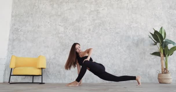 Junge Athletin Tänzerin trainiert zu Hause vor Wandkulisse von Möbeln aus Zimmerpflanzen und gelbem Stuhl dehnt Muskeln der Beine sitzt auf Paddel auf dem Boden und macht Yoga-Übungen steht in Pose Asana — Stockvideo