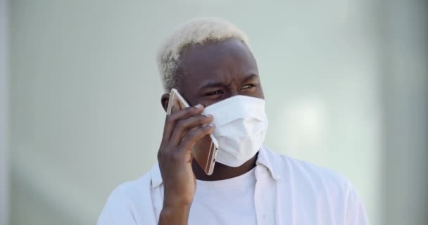 Retrato de un joven hombre afroamericano alegre con camisa casual en máscara médica protectora hablando por teléfono inteligente móvil parado al aire libre. Concepto de protección de la salud pandémica del virus Corona — Vídeos de Stock