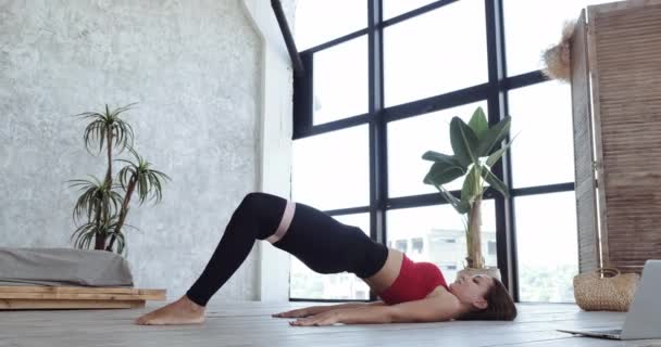 Mulher esportiva treinando nádegas com banda elástica em casa quarto dentro de casa. Mulher fitness fazendo exercícios de treino com elástico deitado no chão olhando na tela do laptop de vídeo on-line com treinador — Vídeo de Stock