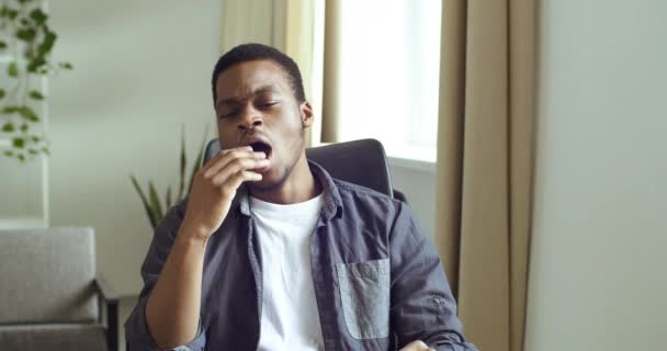 Portrait of Afro American guy in casual clothes sitting at table at home or office feeling bored. Tired man yawns opens his mouth covering his hand suffers from insomnia lack of energy needs coffee — Stock Video