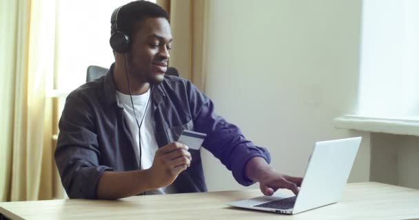 African man freelancer student consumer buyer sitting at home table wears headphones listening to music holds credit card in hands pays online in network makes internet purchase, sale discount concept — Stock Video