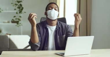 Afro-Amerikalı genç serbest çalışan koruyucu maske takan bir ofis çalışanı evinde laptopun önünde meditasyon yapıyor. Rahatlık hissediyor stresten kurtuluyor. Huzur içinde nefes alma egzersizleri yapıyor.