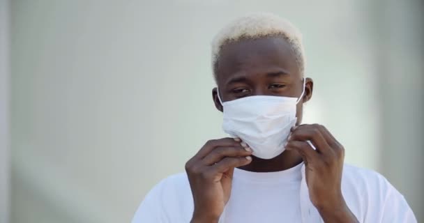 Zbliżenie mieszanej rasy męskiej twarzy. Portret afrykańskiego mężczyzny ubranego w białe, luźne ubrania, noszącego maskę medyczną, chroni zdrowie przed infekcją, zapobiega pandemii wywołanej wirusem Covid. — Wideo stockowe