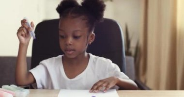 6-7 yaşlarındaki tatlı afro kız ilkokul çocuğunun portresi evde oturuyor ve kağıt üzerinde resim yapıyor. Renkli kalemler kullanıyor. Karışık ırk odaklı çocuklar resim çizer