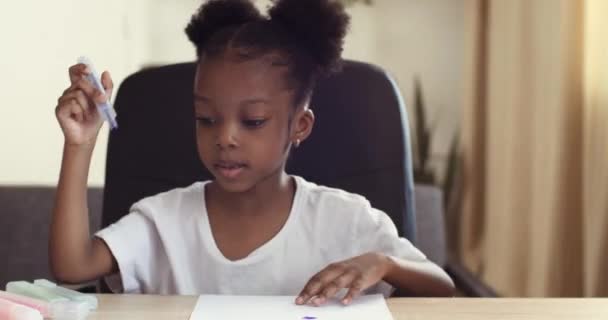 Portrait of happy little afro girl cute primary child school kid of 6-7 years old at home relaxing sits in house and paints on paper use color pencils. Mixed race baby focused children draws picture — Stock Video