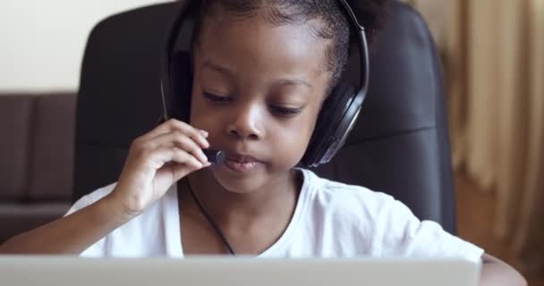 Niña africana juega en el ordenador portátil niño finge administrador de llamadas, colegiala hija lleva auriculares en la cabeza y escucha música mientras está sentado en la mesa de casa, bebé apasionado por los juegos de ordenador — Vídeo de stock