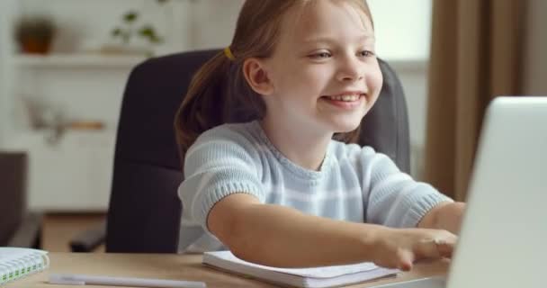 Roztomilá malá školačka dcera s vzrušenými výrazy obličeje hrát online hry na notebooku psaní klávesnice sedí u stolu. Dítě se vzdáleně učí v konceptu karantény, domácí výuky — Stock video