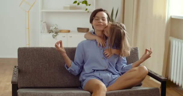 Happy healthy mother doing yoga exercise sit in lotus pose on sofa coupe with close eyes, active kid daughter cute child girl hugs kiss mom distracts from practice naughty baby, loving family concept — Stok Video