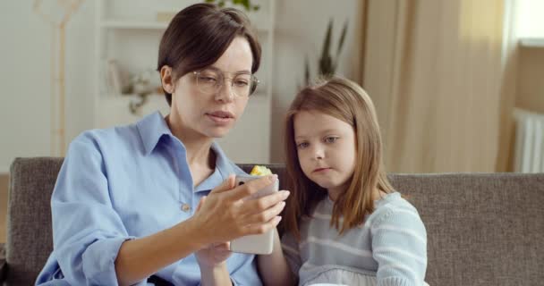 Junge Mutter mit Brille Schwester Nanny sitzt mit ihrer Tochter Mädchen Blick auf Telefon zusammen mit Smartphone machen Foto oder Video. Kinderfrau, die Apfel isst, umarmt Mutter schaut auf Bildschirm ihres Mobilgeräts — Stockvideo