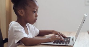 Afro-Amerikalı anaokulu kızı evdeki masada oturan dizüstü bilgisayarın klavyesini daktilo ediyor. Çocuk bilgisayar ekranına odaklanmış. Çevrimiçi öğrenme, mesafe dersi, evde eğitim, çocuklar için teknoloji