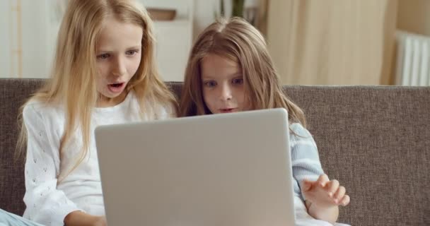 두 명의 행복 한 귀여운 자매 아이들이 원격 교육을 위해 노트북 컴퓨터를 이용하여 소파에 앉아 즐겁게 놀거나 공부하고 있습니다. 작은 코카서스 소녀들이 인터넷으로 재미를 본다 — 비디오