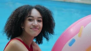 Siyah saçlı Afro-Amerikalı genç kız kameraya bakıp gülümsüyor, havaya plastik şişirilebilir donut tüpü fırlatıyor, çemberle oynuyor, yaz ve tatilin tadını çıkarıyor, havuzda vakit geçiriyor, manzarayı kapatıyor.
