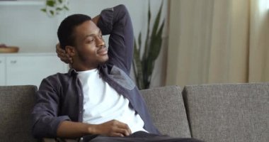 Sakin, düşünceli, Afro-Amerikan adam. Tişörtünü giy, dinlen. Rahat bir evde, kanepede uzanan, elleri bağlı, bilinci açık, genç, melez, kanepede uzanan siyah adam.