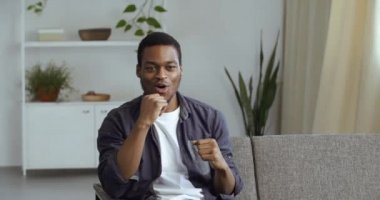 Genç hevesli Afrikalı adam oturma odasında kanepede oturmuş TV seyrediyor çevrimiçi şampiyonluk maçı izliyor boksörleri destekliyor hareketlerini tekrarlıyor havadaymış gibi davranıp el çığlıkları atıyor.