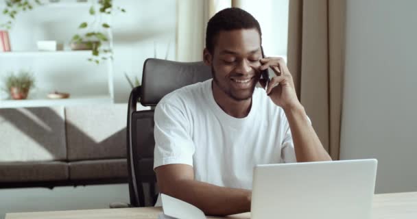 Портрет афро-американского студента-бизнесмена, разговаривающего по мобильному смартфону с клиентом, смотрящим на экран ноутбука, говорящего о стартапе, работающем с домашнего пульта — стоковое видео