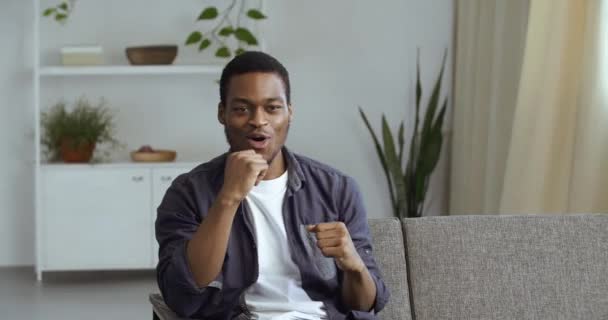 Joven chico africano entusiasta sentado en casa en la sala de estar en el sofá viendo la televisión campeonato en línea apoya boxeador repite movimientos fingiendo caja en el aire con las manos grita grita con deleite — Vídeo de stock