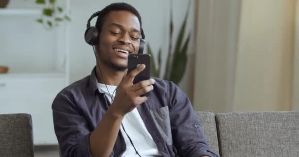 Hombre de raza mixta afroamericano en ropa casual se sienta en casa sala de estar en el sofá tiene teléfono inteligente en sus manos lleva auriculares escuchando música en línea en la radio bailando activamente en movimiento divertirse — Vídeo de stock