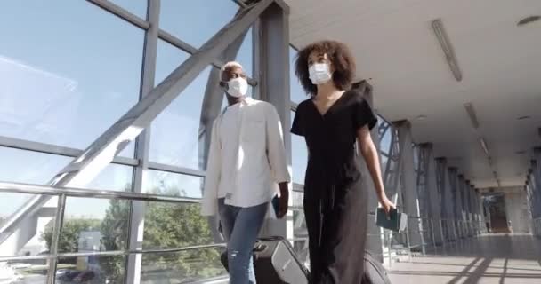 Kilku afrykańskich nastolatków przyjaciół nowożeńców mężczyzna i kobieta w maskach medycznych spacerujących po terminalu lotniska niosących walizkę z bagażem trzymającym bilety lotnicze lądujące paszport rozmawiający ze sobą — Wideo stockowe