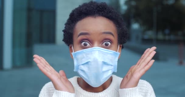 Närbild av överraskad kvinna av afrikansk etnicitet bär medicinsk mask på kvinnligt ansikte aktivt gestikulerar med händerna känns chock förvåning säger wow känsla av skräck står utomhus nära byggnaden — Stockvideo