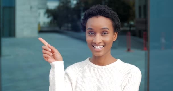 Ritratto di giovane donna afroamericana sorridente adolescente ragazza studentessa in stand bianchi posa guardando la fotocamera all'aperto punta dito a lato mostra la direzione fa la scelta a mano, primo piano — Video Stock