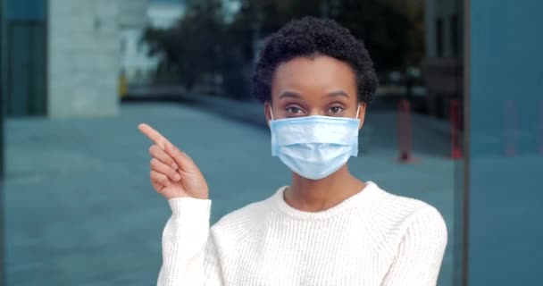 Close-up retrato africano mestiço mulher menina fêmea paciente usa máscara cirúrgica protetora médica em face aponta dedo para hospital fica ao ar livre isolado um à distância olhando para a câmera — Vídeo de Stock