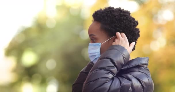 秋の天候で屋外でカメラに向かって横に立っているアフリカの女性の肖像は、パンデミックの間にコロナウイルス感染を防ぐために彼女の顔の上に医療保護マスクを置きます。女性のプロフィール閉じる — ストック動画