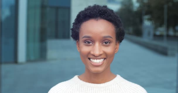 Portret van mooie gelukkige verlaten stijlvolle Afrikaans Amerikaanse vrouw student meisje vrouwelijk model tanden glimlachen op camera kijken zelfverzekerd dragen van witte kleren poseren op stedelijke stad achtergrond, close-up — Stockvideo