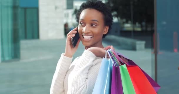 Afro-Amerikalı mutlu bir kadının portresi gülümsüyor. Sokakta gülüyor, alışveriş paketleri elinde telefonla konuşuyor. Arkadaşına indirimlerden bahsediyor. — Stok video