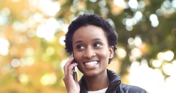 Portré afrikai etnikai nő beszél telefonon mosolygós fogú, használja a modern okostelefon eszköz válaszolni hívás kommunikáció barátaival, nevetve áll a szabadban ősszel város, visel meleg kabát — Stock videók