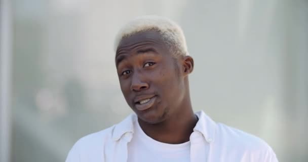 Πορτρέτο του συναισθηματικά ενεργό χαρούμενος τύπος αφροαμερικανός έφηβος φοιτητής στέκεται σε εξωτερικούς χώρους με λευκά ρούχα χαμόγελα toothy κοιτάζει κάμερα φιλικό μιλάει χαιρετισμό χαιρετισμό χέρι προς τον εαυτό του — Αρχείο Βίντεο