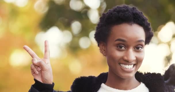 Attivo felice giovane donna africana studentessa straniera ragazza di successo soddisfatta signora si trova da solo di fronte alla fotocamera all'aperto vicino albero autunnale mostra segno di pace con due dita, simbolo di sostegno amichevole — Video Stock
