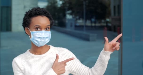 Mulher africana em stands máscara mostra com as mãos direção a lado. Paciente de raça mista usa equipamento de proteção médica no rosto vira os dedos para a direita explica em linguagem corporal onde é hospital — Vídeo de Stock