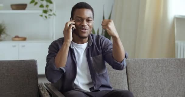 Afro student learns good news on phone enjoys sitting at home. Black male businessman freelancer speaks by smartphone celebrating triumph. Male consumer answers call hears about discount feels success — Stock Video