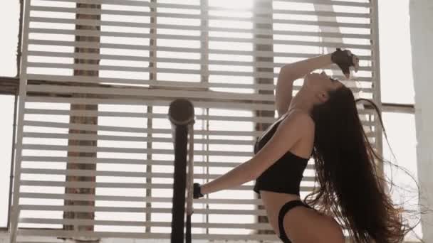Charmant modèle de fitness brunette femme en noir sujet peluches cheveux longs avec la main appuyée sur la rampe dans le studio de gym, fond de soleil de la fenêtre avec volets. matin lumineux de belle fille athlétique — Video