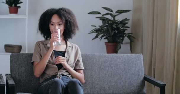 Красивая смешанная раса девушка подросток сидит расслабляется на диване в квартире после трудного дня, освежается холодной минеральной водой, держит в руках стекло, пьет вкусный напиток, здоровое питание концепции — стоковое видео