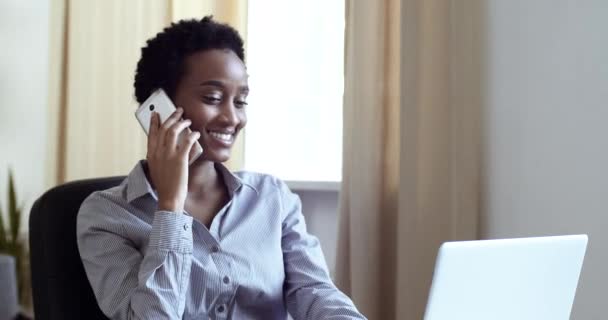 Serious atraente afro-americana empresária estudante menina freelancer falando ao telefone com colegas clientes sentados à mesa no local de trabalho em frente ao laptop consulta remotamente a partir de home office — Vídeo de Stock