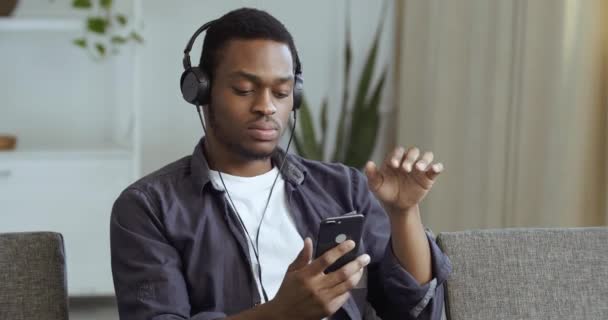 Homem negro calmo afro-americano sentado em casa no sofá na sala de estar isolado em quarentena ouve música em fones de ouvido detém smartphone em suas mãos finge tocar piano com os dedos — Vídeo de Stock