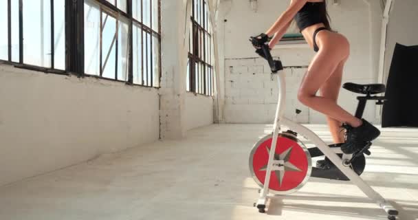 Hermosa atlética modelo de fitness de mujer joven en tanga negra y zapatillas de deporte que entrenan activamente en bicicleta estática en el gimnasio blanco casero aislado cerca de grandes ventanas bajo el sol. Chica quema calorías en simulador. — Vídeo de stock