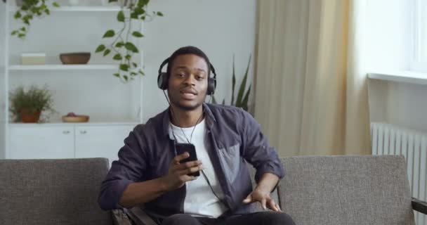 Hombre afroamericano escuchando música sentado en el sofá en la sala de estar en casa. negro chico estudiante descansando en sofá usa auriculares utiliza teléfono inteligente disfruta en línea radio canta canciones mueve sus manos danzas — Vídeos de Stock