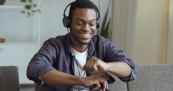 Ativo cara feliz afro-americano adolescente negro usa fones de ouvido segurando smartphone móvel em mãos ouvindo música áudio on-line rádio hip hop em casa descansando no sábado dançando sentado no sofá — Vídeo de Stock