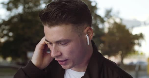 Portré fiatal kaukázusi diák vidám férfi vezeték nélküli fejhallgató fülébe, amelynek telefonhívás és aktívan beszél mosolyogva áll a szabadban. Stílusos srác beszélni okostelefon a kommunikációhoz — Stock videók
