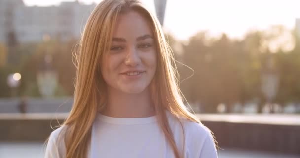 Venku portrét mladé blondýny dívka teenager blondýny mluví při pohledu na kameru usmívá přátelsky narovnává její dlouhé vlasy, atraktivní žena pózuje na ulici ve větrném počasí, mládež krása koncept — Stock video