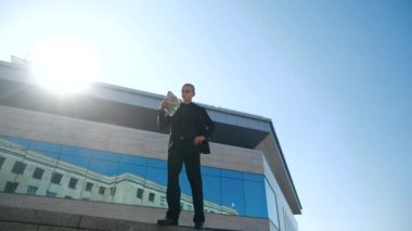 İşadamı yönetici takım elbise giyer, açık havada modern mavi cam binanın yanında durur, ellerinde dolarları tutar, yüzüne para dalgaları savurur, kendine güvenen işadamı başarıyı hisseder, maaşını alır.