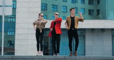 Parlak gömlekli üç yakışıklı adam dans hareketlerini prova ediyorlar, aynı anda kollarını yana doğru hareket ettiriyorlar, dirseklerini büküyorlar, açık havada dans ediyorlar, performanslarını gösteriyorlar.