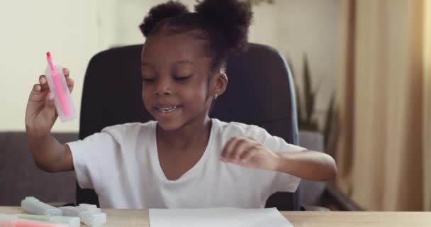 Portrait of little black schoolgirl child sits at home alone draws with bright colored markers with felt-tip pens pencils on paper picture studying does homework comments tells explains, close up — Stock Video